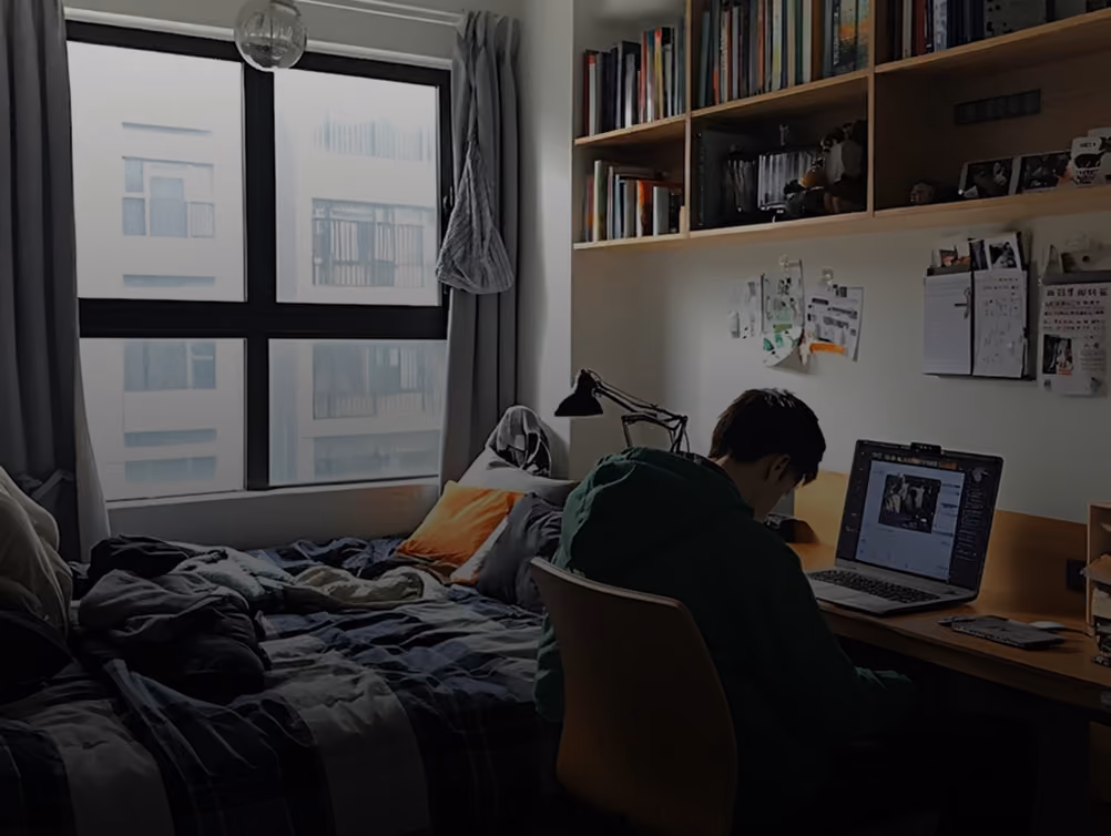 Student studying on a laptop in a dorm room with a bed, window, and bookshelves.
