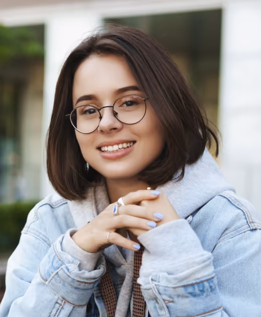 Smiling young woman with glasses wearing a denim jacket and gray hoodie outdoors.