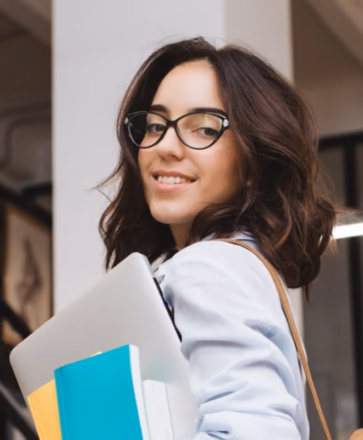 Smiling woman with glasses holding books and a laptop, looking back over her shoulder indoors.