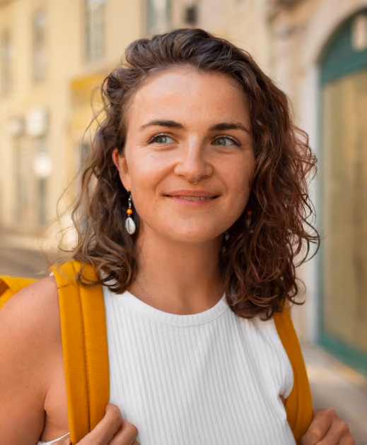 Smiling woman with curly brown hair wearing white top and mustard yellow backpack outside on a street.