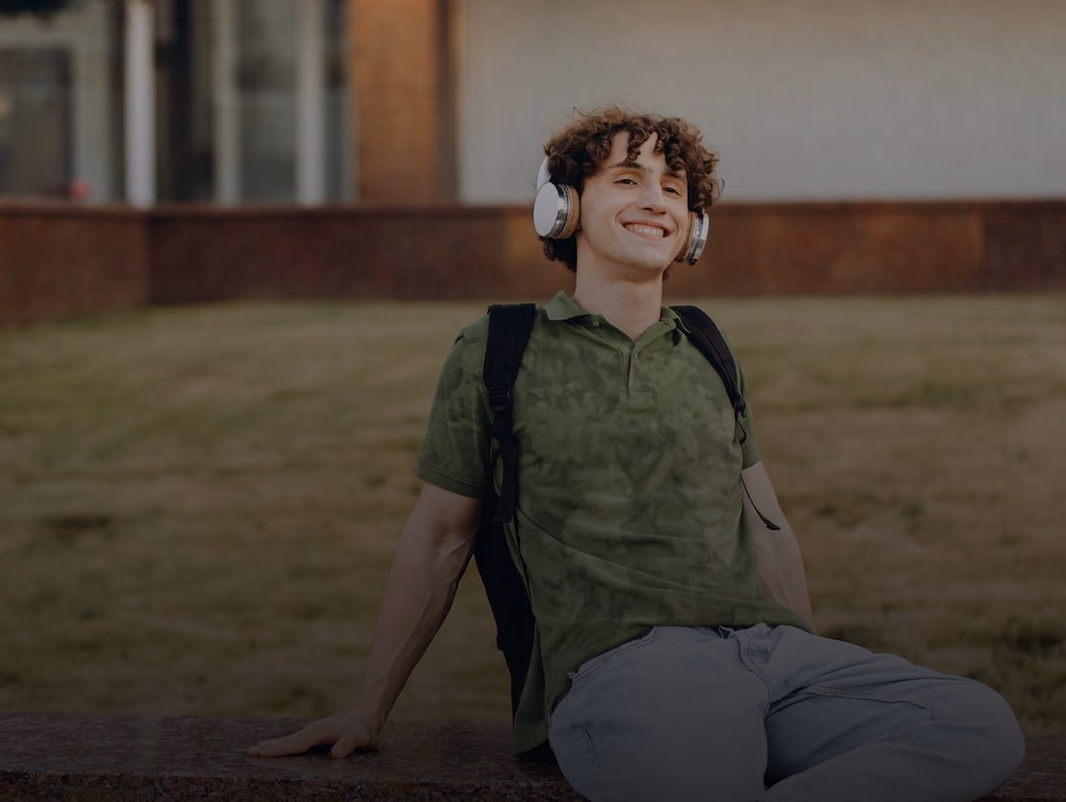 Smiling young man with curly hair wearing headphones, green shirt, and backpack, sitting outdoors.